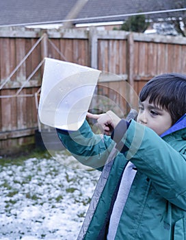 Boy holding measuring jug pointing at level of rain collected in garden. 6 year old child measuring rainfall for school science