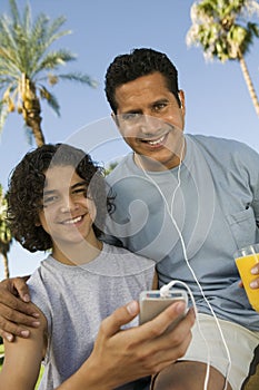 Boy (13-15) holding portable music player father listening with earphones and holding glass of juice front view portrait.