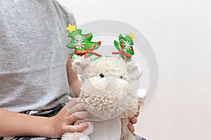 A boy holding a soft toy, teddy hippo with christmas holiday accessories, carnival costume with toy fir trees on white background