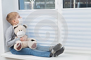 Boy holding teddy bear in protective mask loking through the window