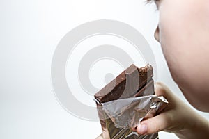 The boy holds a bar of chocolate and eats on a white background.