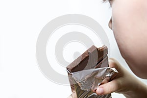 The boy holds a bar of chocolate and eats on a white background.