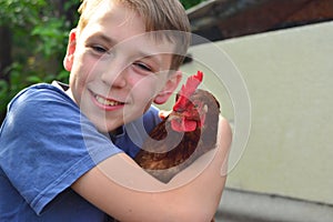 A boy holds a brown chicken in his hand and smiles and kisses her and hugs him.