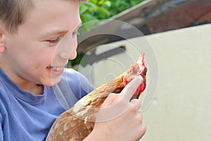 The boy holds a brown chicken in his hands and smiles and kisses her and hugs and holds a finger for his beak