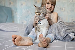 boy holds a cute domestic cat between his bare feet while sitting at home in the morning on the bed