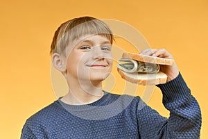 The boy holds in his hand a sandwich with bread and dollars near his open mouth
