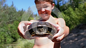 Boy Holds Turtle in Arms and Smiles Viciously on River with Green Vegetation