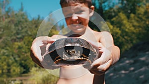 Boy Holds Turtle in Arms and Smiles Viciously on River with Green Vegetation