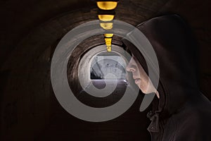 Boy with hood standing in a dark underground passage with yellow light bulbs at the roof and light at the end. Black and white pho