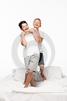 Boy jokingly stifling smiling brother while standing on bed isolated on white