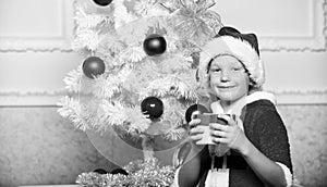 Boy kid dressed as santa with red hat hold gift box near christmas tree. Christmas santa claus costume for child