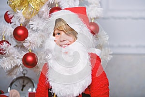 Boy kid dressed as santa with white artificial beard and red hat near christmas tree. Christmas santa claus costume for