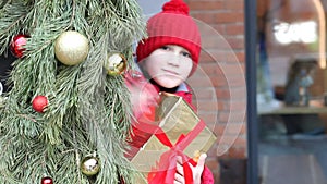 Boy kid in winter clothes standing outside with decorated Christmas tree, holding gift box and waving hand