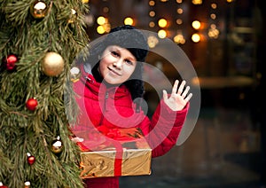 Boy kid in winter clothes standing outside with decorated Christmas tree, holding gift box and waving hand