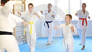 Boy in kimono performing punch moves in gym during group training with trainer, in background his family is doing kata