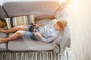 Boy laying on sofa plays with electronic devices - gamepad connected with smartphone