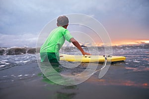 Boy learning to surf in ocean waves at sunset time