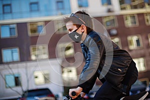 Boy in a leather jacket wearing a black hygyenic medical mask with white cross outdoors in the yard, riding a bike