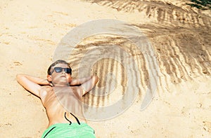 Boy lies on sand in palm tree shadow