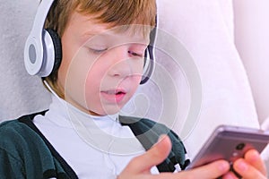 Boy is listening music in headphones in smartphone and singing a song sitting in armchair at home.