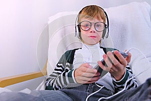 Boy is listening music in headphones in smartphone and singing a song sitting in armchair at home.