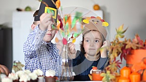 Boy and little cute girl in paper turkey and pilgrim hat glues paper leaves on branches for celebrating Thanksgiving day