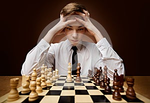 Boy looking at Chess Board Checkmate holding Head in Hands. Stressed Teenager in Business Shirt thinking about Strategy
