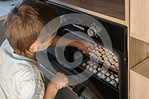 Boy looks into opened oven on white cooking meringue and picks up one with his hand.