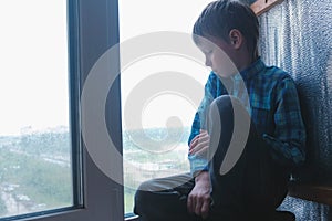 Boy looks out the window sitting in windowsill in the rain and is sad.