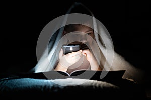 A boy lying in bed reading a book in the dark, illuminating with a flashlight from the phone