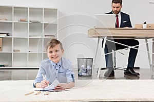 Boy lying on carpet and drawing while his father businessman working