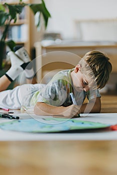 Boy lying on stomach and painting at home with watercolors and tempera paints, creating a model of planet Earth.