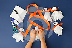 Boy makes Halloween toys ghost from white paper napkin. Creative DIY for kids on classic blue background. Home decor project party