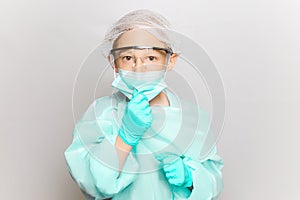 Boy in a medical gown and mask adjusts the mask with his hand which is wearing disposable gloves