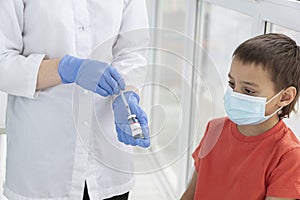 Boy in medical mask is lookig at doctor`s hands with vaccine and syringe, doctor prepares to vaccinate young patient.