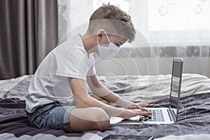 A boy in a medical mask sits in front of a laptop at home in bed. Distance learning