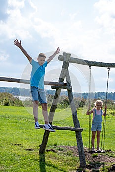 A boy is mid air and jumping from a play structure as a girl swings nearby, capturing a moment of youthful exuberance