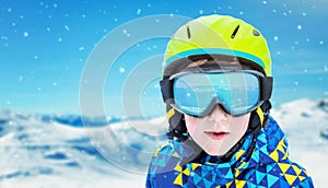 Boy with modern ski green helmet, yellow glasses and blue jacket. Reflection of snowy peaks on glasses