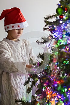 A boy in a New Year's hat is decorating a Christmas tree at home