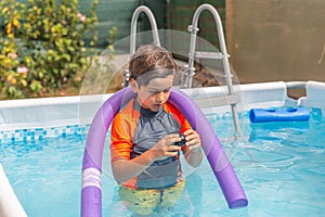 Boy in orange swim shirt plays with a large purple noodle in a sunny home swimming pool setting