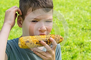 A boy outdoors on a background of green grass eats yellow corn and thinks of something, in front there is a place for inscription
