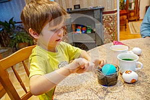 Boy paints eggs for Easter, use spoon dips eggs into colored water in the home interior