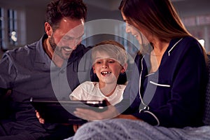 Boy With Parents In Pyjamas Sitting On Sofa Watching Digital Tablet Together