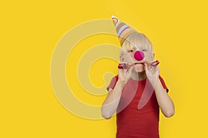 Boy with party hat and red clown nose holding whistle. Portrait on yellow background. Holiday matinee