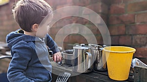 A boy playing with a mud outdoor kitchen holding utensils