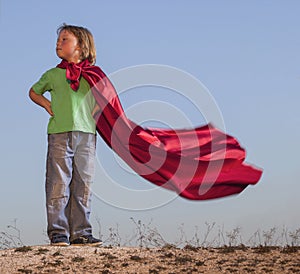 Boy playing superheroes on the sky background, superhero in a red cloak on hill