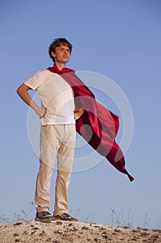 Boy playing superheroes on the sky background, teenage superhero in a red cloak on hill