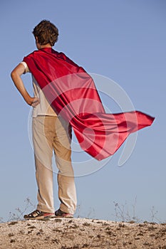 Boy playing superheroes on the sky background, teenage superhero in a red cloak on hill
