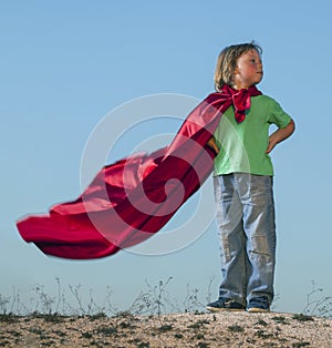 Boy playing superheroes on the sky background, teenage superhero in a red cloak on hill
