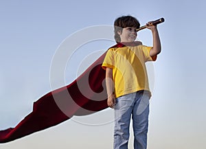 Boy playing superheroes with spyglass on the sky background, teenage superhero in a red cloak on a hill